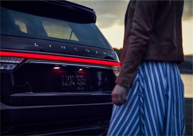 A person is shown near the rear of a 2023 Lincoln Aviator® SUV as the Lincoln Embrace illuminates the rear lights | South Bay Lincoln in Hawthorne CA