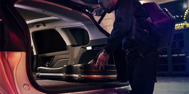 A person loading luggage into the open rear liftgate of a three-row SUV with spacious cargo area and folded third row.